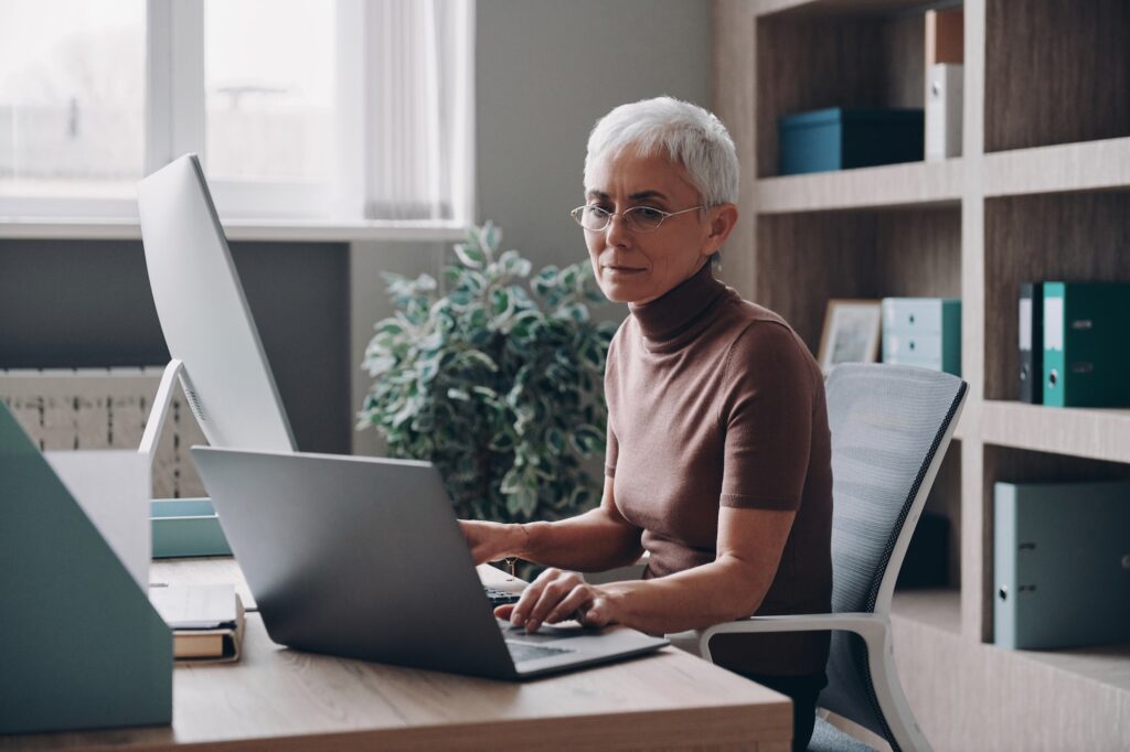 Confident senior businesswoman working at two computers while sitting in the office