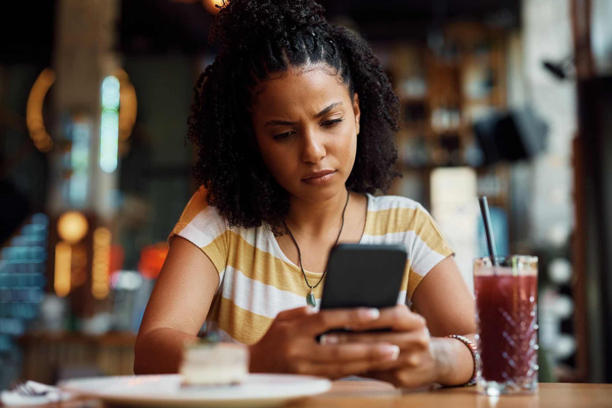 Distraught African American woman reading text message on mobile phone in a cafe.