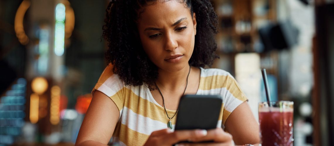 Distraught African American woman reading text message on mobile phone in a cafe.